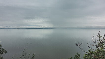 Serene seascape. A lone ship is visible on the surface of the Pacific Ocean. Cloudy sky, fog over the water. Green vegetation in the foreground. Kamchatka