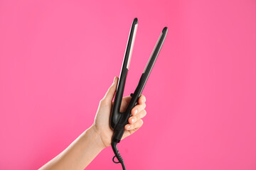 Woman holding flat hair iron on pink background, closeup