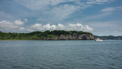A white yacht sails along the picturesque coast of Kamchatka. Rocks and green hills against a background of blue sky and clouds. Ripples on the water of the Pacific Ocean. Avacha Bay