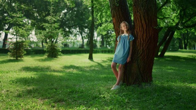 Dreamy girl lean tree trunk in park. Smiling kid observing nature around