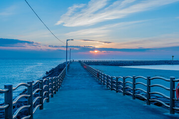 Obraz premium Long concrete pier at seaport with setting sun behind clouds in background.