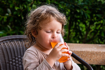 a little girl with curly hair drinks an orange drink from a tall glass glass in summer. the child drinks orange juice. quench your thirst in summer. colored drinks