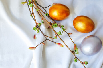 Easter eggs of golden and silver color lie on a white tablecloth, next to a branch with buds.