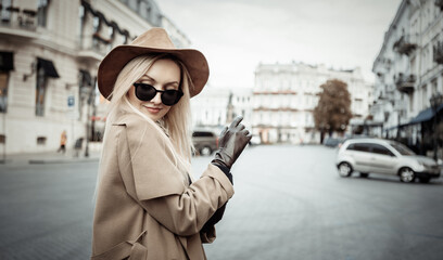 Portrait of young stylish blonde woman in autumn coat, sunglasses and felt hat wear gloves in...