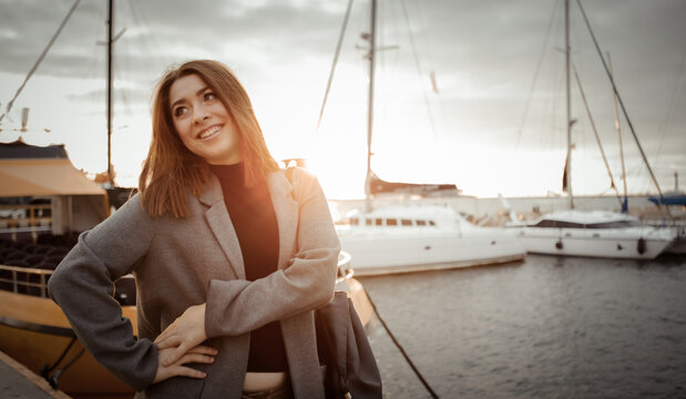 Portrait of a young cheerful woman in an autumn coat posing in a yacht club at sunrise