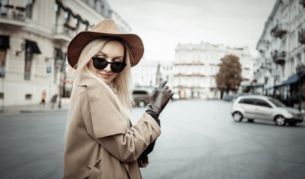 Portrait of young stylish blonde woman in autumn coat, sunglasses and felt hat wear gloves in European city. Beautiful attractive girl