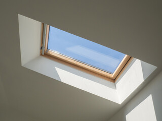 Dormer wooden window in the white sloping ceiling overlooking the blue sky. Sunlight enters the...