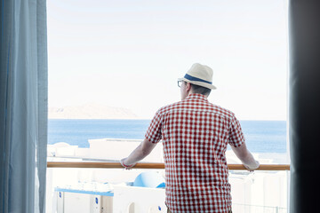 Handsome Caucasian young man standing on balcony of hotel, looking at sea view