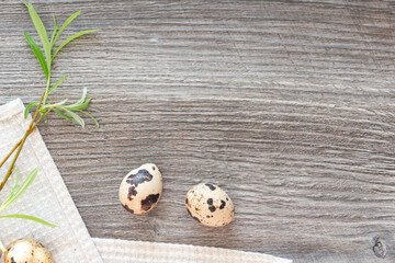 Quail eggs and a green willow branch are on a linen napkin, on a gray and wooden table.