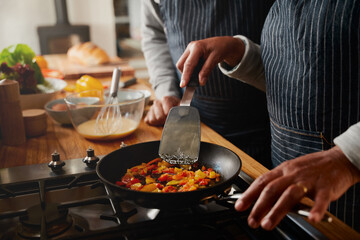 Multi-cultural elderly couple frying vegetables with spatula in pan at modern stove.