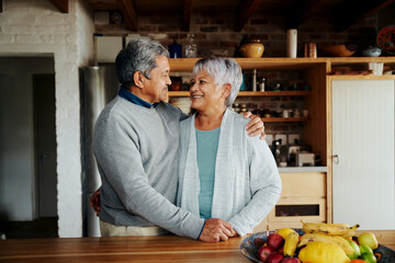 Happily retired elderly biracial couple standing, hugging and smiling at each other in kitchen.