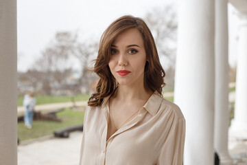 pretty caucasian young woman in beige blouse with seductive red lipstick and wavy long hair standing on terrace with white concrete pillars