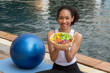 black woman sitting by the pool on a yoga mat having a blue yoga ball, finishes her workout, eats...