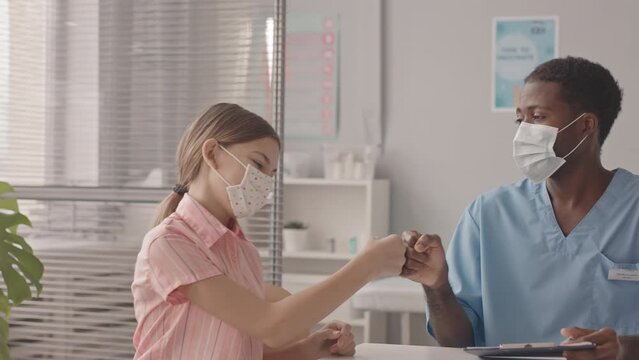 Medium slowmo shot of 13-year-old Caucasian girl in face mask having medical appointment at doctors office, greeting her African-American pediatrician with fist bump, preparing for vaccination