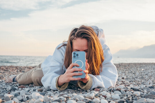 Happy young woman lying on pebble beach and taking selfie portrait at sunset during golden hour. 