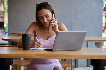 young asian pretty woman in pink dress sitting outdoors, writing notes in notebook, calling by smartphone. attractive female student with cup of coffee at cafe studying. lifestyle, urban portrait