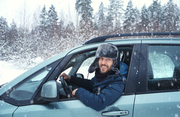 Happy positive man driver looking on camera thowth window of his car enjoying snowy day on country road.  Road trip, winter vacation. Driving auto in cold season.