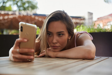 young asian pretty woman with long hair lays her head on her arms and table, holding phone in hand....