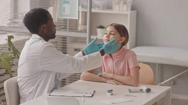 Medium slowmo shot of 13-year-old Caucasian schoolgirl at medical check-up having her lymph nodes examined by young African-American doctor in lab coat