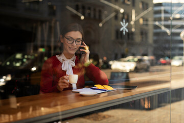 Stylish young woman drinking coffee at cafe. Beautiful girl talking to the phone while enjoy in fresh coffee.
