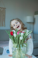 Cute little girl of 3 years old in a cozy knitted sweater hugs a vase with a bouquet of white and pink tulips in a home interior. Home comfort. Spring.