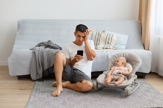 Image of tired exhausted man wearing white casual style T-shirt and jeans short sitting on floor with newborn baby, father didn't sleep whole night, posing closed eyes.