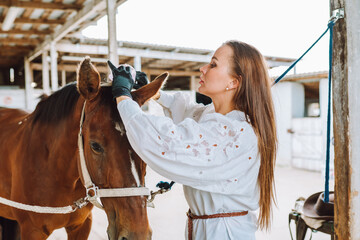 Beautiful brunette horsewoman equestrienne put on harness. Horse back riding. Cleaning horse shoe, mane. Animal care. Bound between owner and horse. Outdoor farm, paddock, yard. Countryside. Side view