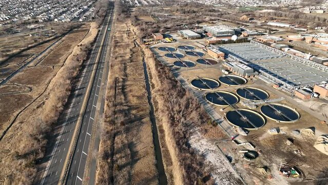 A high angle aerial view of a waste water treatment plant on a sunny day. The camera tilt down, dolly in between the long road and surrounding marsh land and the large processing plant.