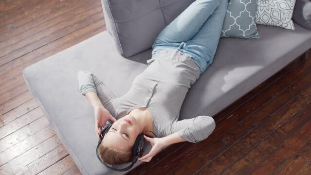 Young beautiful girl lies on a sofa in headphones and listens to music. Blond woman is resting at home.