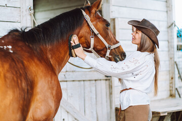 Tender horsewoman equestrienne in hat, trousers, shirt, belt. Cleaning, grooming horse mane. Animal care. Horse back riding. Bound between owner and horse. Outdoor farm, stall, stable. Side view