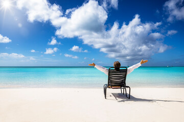 A happy man relaxes in a chair on a tropical paradise beach in the Caribbean with turquoise sea and blue sky during his vacation time - obrazy, fototapety, plakaty