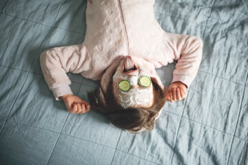 Cute funny caucasian european girl child in soft pajamas with a mask on her face and cucumbers on her eyes. humorous concept of self care and cosmetology. Toning and lifestyle in a real interior
