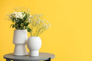 Vases with roses and gypsophila flowers on table near yellow wall