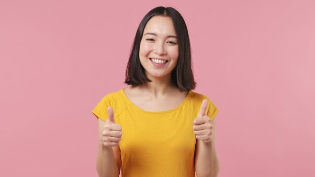 Excited cheerful young woman of Asian ethnicity 20s wears yellow t-shirt showing thumb up like gesture isolated on plain pastel light pink background studio portrait. People emotions lifestyle concept