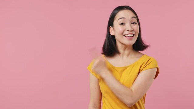 Promoter young woman of Asian ethnicity 20s in yellow t-shirt pointing fingers aside on workspace copy space mockup promo commercial area isolated on plain pastel light pink background studio portrait