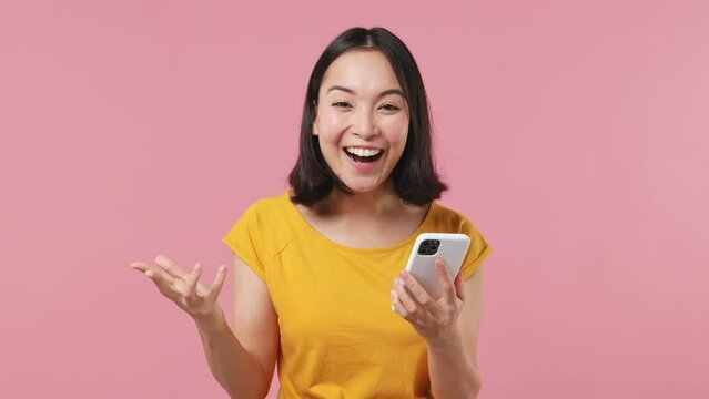 Surprised joyful young woman of Asian ethnicity 20s in yellow t-shirt hold use mobile cell phone typing say wow yes doing winner gesture isolated on plain pastel light pink background studio portrait