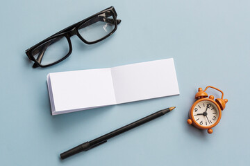 Notepad with reading glasses, pen and alarm clock on blue background