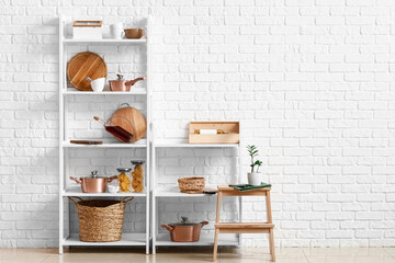 Fototapeta na wymiar Modern shelf units with kitchenware and stand near white brick wall