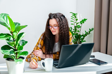 Smiling woman in glasses writing in a notebook on a workplace. Woman working at home, takes notes at a meeting, undergoing training, watching webinar