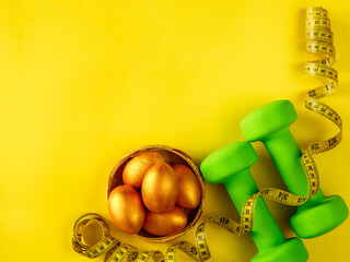 green heavy dumbbells, golden easter eggs and yellow tape measure on yellow background. Easter nutrition, fitness and training composition with copy space. Top view from above, flat lay