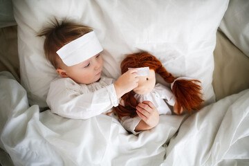 Cute baby with a doll with a compress on his head, a sick child in bed, bed rest during illness. Comfort and play in bed