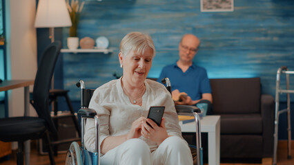 Senior person sitting in wheelchair and using smartphone to browse internet and discover...