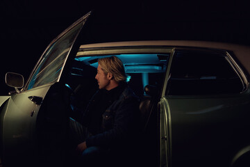 Man with blond hair sits inside an American classic muscle car with opened door at night.