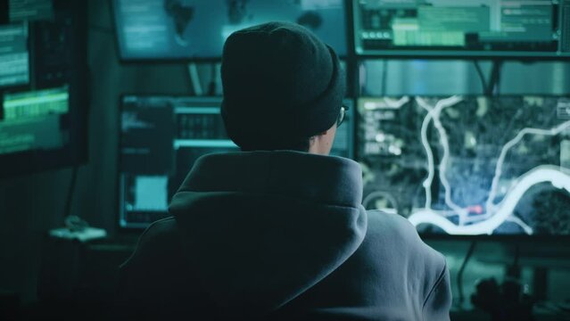 Back view of young man in hoodie and hat typing on computer keyboard and spying on victim while sitting at table in dark room
