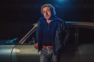 Man with blond hair in a jeans jacket stands by an American classic muscle car.
