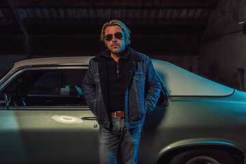 Man with blond hair and sunglasses in a jeans jacket stands by an American classic muscle car.