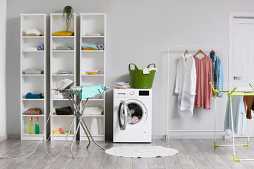 Interior of light laundry room with shelf units, washing machine and rack with clothes
