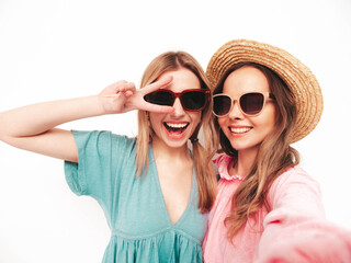Two young beautiful smiling brunette hipster female in trendy summer dresses. Sexy carefree women posing near pink wall. Positive models having fun.Taking Pov selfie. In hats and sunglasses