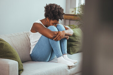 Frustrated young lady sitting on sofa, cuddling pillow, looking away at window. Lost in thoughts...