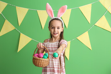 Little girl with bunny ears holding basket of Easter eggs on green background